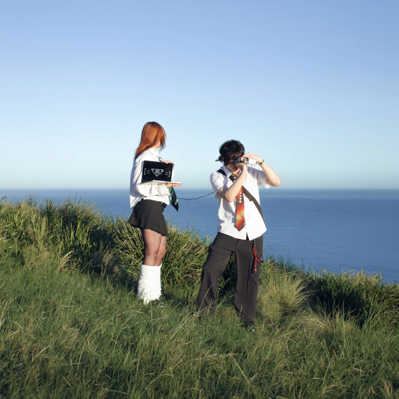 Ninajirachi and Porter Robinson single artwork for WannaCry featuring the duo on a grassy ocean cliff with a laptop and camcorder under clear sky.
